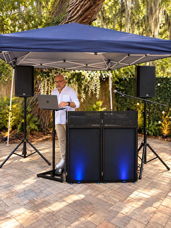 A clean DJ facade, two speakers and a microphone prepared beneath a canopy for a garden event.