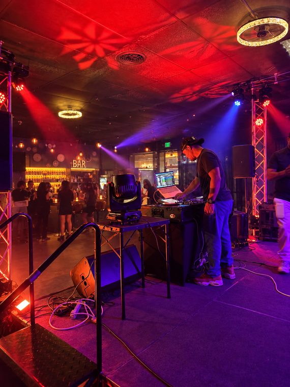 A DJ performing on a club stage surrounded by moving lights, truss and professional sound equipment.