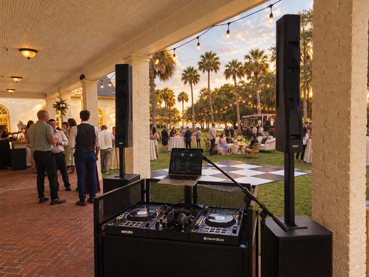 A DJ booth and two column-array systems overlooking a dance floor and outdoor reception at sunset.